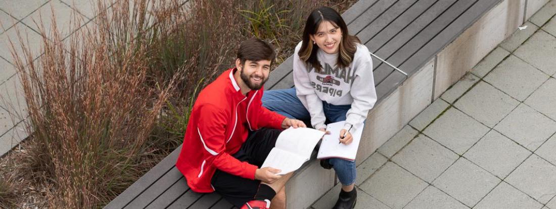两个新葡新京娱乐场app下载的学生拿着书坐在校园外面，对着镜头微笑